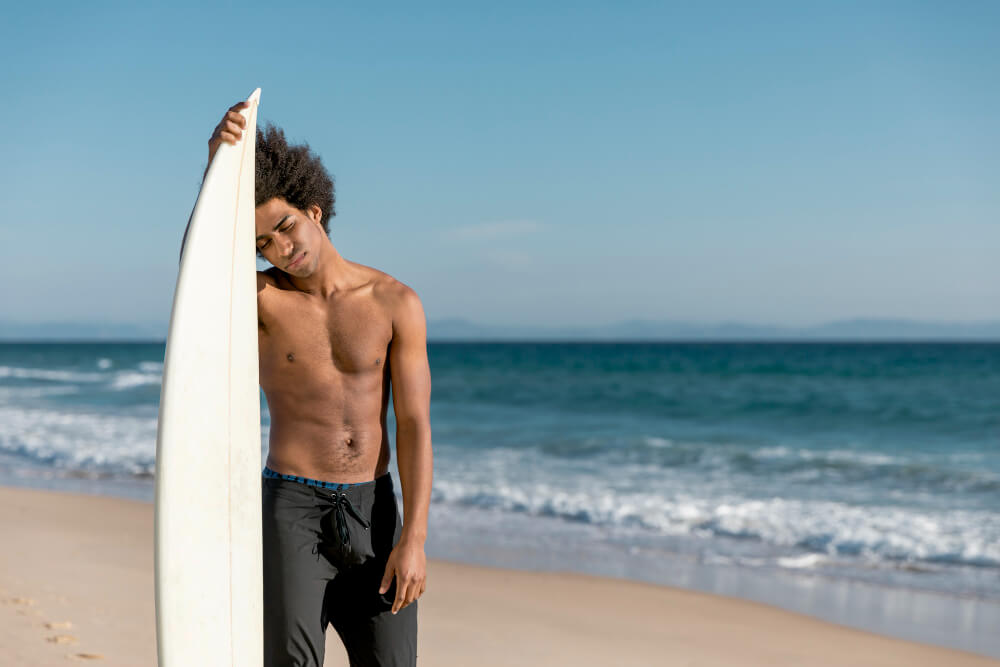 A tired-looking surfer, standing on a beach with a pained expression on his face