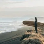 A lone surfer stood on top of a dune, staring at the sea