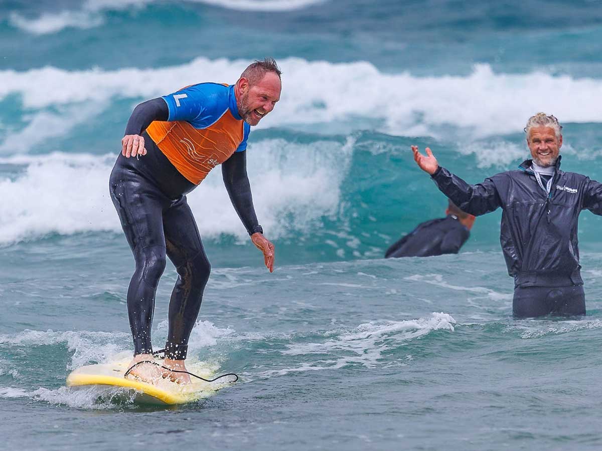Head Coach Julian cheering on an older man who is riding a surfboard standing up