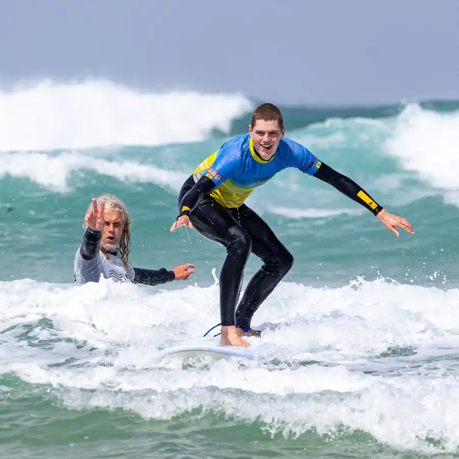 A smiling man riding a wave on a surfboard, ith head coach Ju standing in the sea behind him
